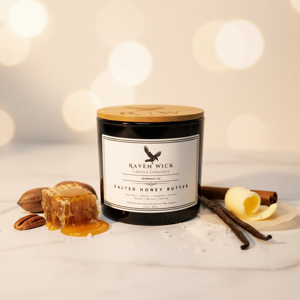 A black glass jar candle with a light wood lid and a white label reading Raven Wick Candle Company, Salted Honey Butter, on a gray surface with a white curtain backdrop.