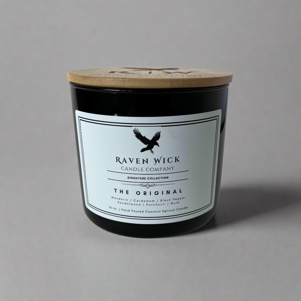 A black glass jar candle with a light wood lid and a white label reading 'Raven Wick Candle Company' and 'The Original', photographed against a plain light gray background.